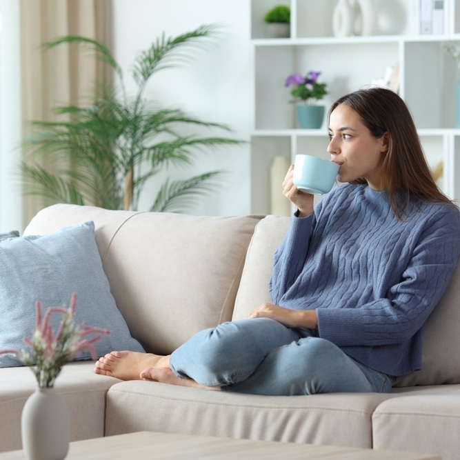 Détente au salon : femme en pull bleu buvant un café Femme en pull bleu se relaxant sur un canapé beige en buvant une boisson chaude dans un salon moderne et lumineux.